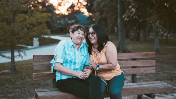 Woman with a peach blouse and jeans on a park bench with a older woman in a blue blouse and jeans facing the challenges of caring for aging parents.