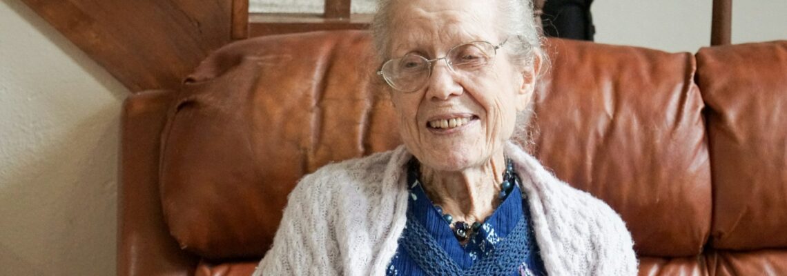 An elderly woman at home on her brown couch smiling an wearing glasses and a white sweater and blue dress aging at home.