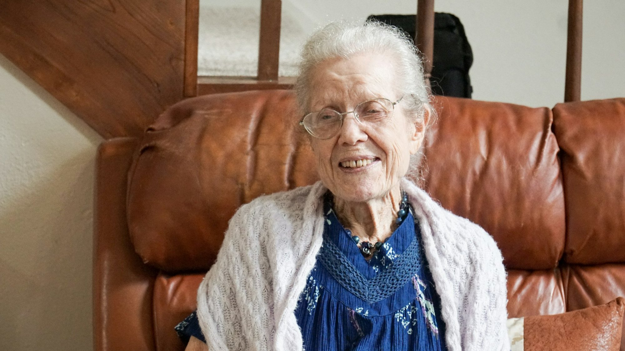 An elderly woman at home on her brown couch smiling an wearing glasses and a white sweater and blue dress aging at home.