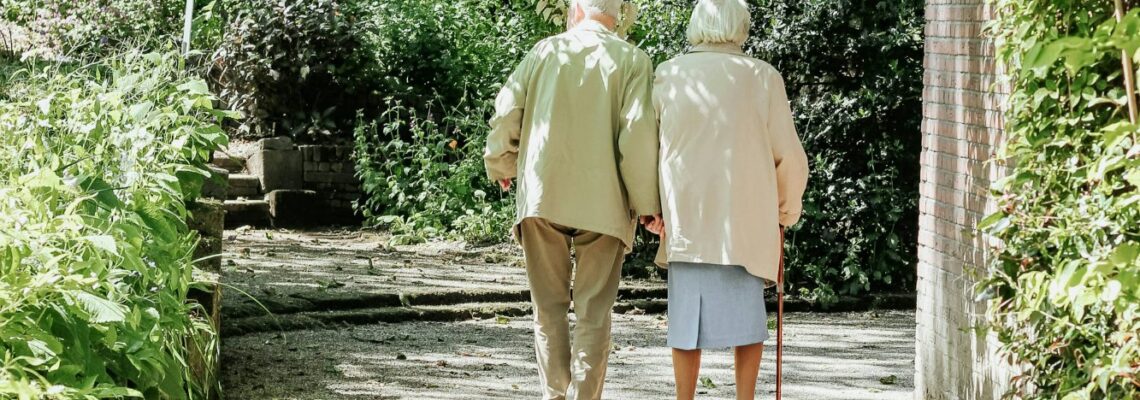 An elderly couple walking in a garden on a sidewalk next to a brick wall exemplifying the need for home care for aging parents.