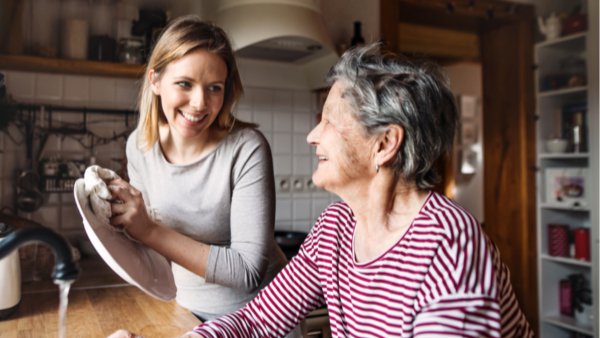 Female Caregiver in gray, long-sleeve blouse washing dishes with elderly woman in a striped shirt providing hone care services.