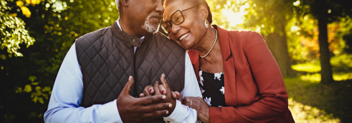 Older couple standing close together outdoors, smiling and holding hands in a sunlit park