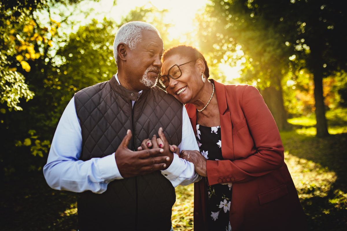 Older couple standing close together outdoors, smiling and holding hands in a sunlit park